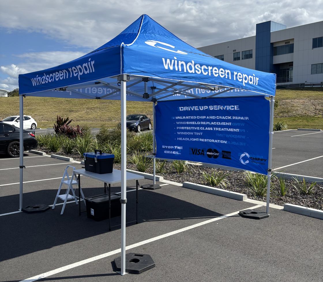 A Chippy Repair kiosk set up in a shopping-centre car park — a branded 3m by 3m marquee standing inside two line-marked parking bays, held down by free-standing ballast weights.